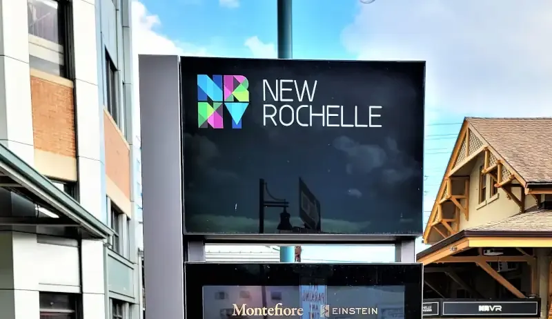 A dark glass pane with colorful font with buildings in background - font reads 'NRNY New Rochelle'